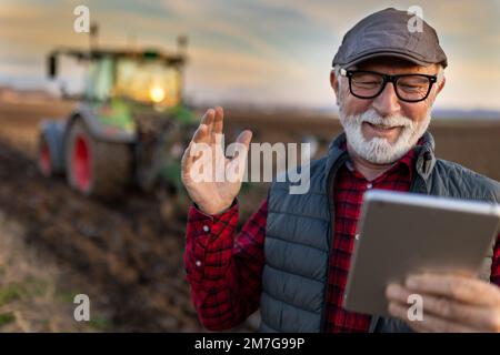 Aufgeregter, reifer Landwirt, der auf dem Feld vor dem Traktor mit einem Tablet arbeitet Stockfoto