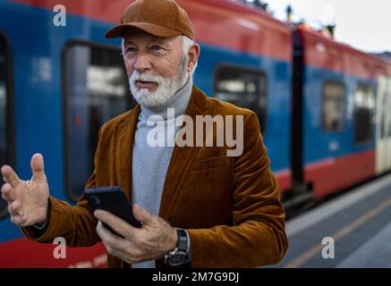 Ein älterer Mann, der vor dem Zug am Bahnhof ein Handy benutzt Stockfoto