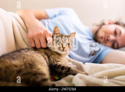 Ein junger Mann, der im Bett mit einer süßen Katzenkatze kuschelt Stockfoto