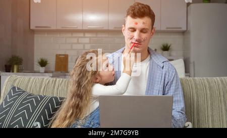 Vorschulmädchen malt Lippen von Vater, der an einem Laptop arbeitet Stockfoto