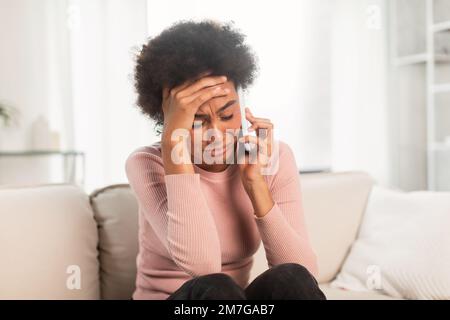 Verzweifelte traurige, deprimierte junge gemischte Rassenfrau, die auf dem Sofa sitzt, per Smartphone anruft, von Hand zu Kopf drückt Stockfoto
