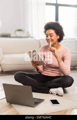 Eine aufmerksam lächelnde junge gemischte Rasse mit Computer macht Notizen, plant Arbeit, sitzt auf dem Boden Stockfoto