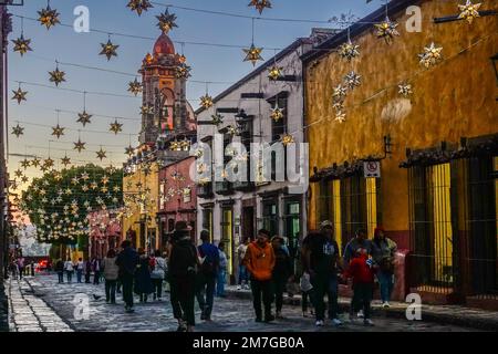Farbenfrohe verzinnte Sterndekorationen auf historischen Gebäuden mit dem Kirchturm der Unbefleckten Empfängnis in der Canal Street in der Abenddämmerung im historischen Viertel, 4. Januar 2023 in San Miguel de Allende, GTO, Mexiko. Stockfoto
