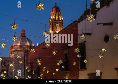 Farbenfrohe verzinnte Sterndekorationen auf historischen Gebäuden mit der Kuppel und dem Kirchturm der Unbefleckten Empfängnis in der Canal Street in der Abenddämmerung im historischen Viertel, 4. Januar 2023 in San Miguel de Allende, GTO, Mexiko. Stockfoto