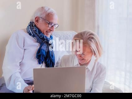 Zwei Großeltern amüsieren sich im Wohnzimmer, während sie auf das Notebook schauen. Sie kommunizieren mit ihrem Enkel. Das Licht ist weich. Stockfoto
