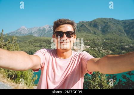 Mann macht Selfie-Foto mit Weitwinkel-Frontkamera mit Blick auf den Hintergrund des Bergsees. Ein männlicher Reisender, der Spaß auf dem blauen See hat, reist im Freien Stockfoto