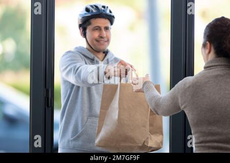 Foto eines Mannes mit Fahrradhelm, der zu Hause ein Lebensmittelpaket mit Fokus auf Hand- und Kopierbereich liefert Stockfoto