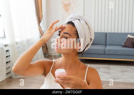 Charmante Brünette Frau mit Handtuch auf ihren Kopf gewickelt Anwendung feuchtigkeitsspendende Gesichtscreme Stockfoto