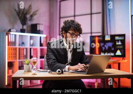 Seitenansicht, Porträt eines jungen, lächelnden, erfahrenen, schlauen, stilvollen, bärtigen Managers mit Brille, Geschäftsmann im Anzug, der am Tisch sitzt und am Abend im modernen Büro einen Laptop benutzt Stockfoto