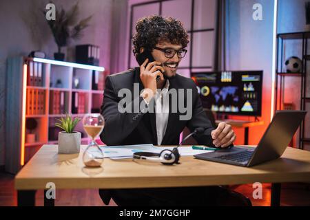 Arbeitsprozess im Büro eines jungen ernsthaften Mannes, der abends mit Partnern telefoniert. Positiver, bärtiger Geschäftsmann, der mobile Gespräche führt, während er am Tisch sitzt und an einem Laptop arbeitet. Stockfoto