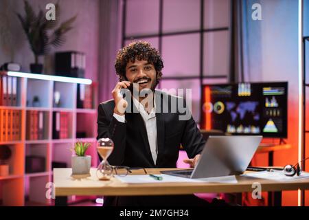 Ein junger Mann, der abends mit Partnern telefoniert und im Büro arbeitet. Positiver, bärtiger Geschäftsmann, der mobile Gespräche führt, während er am Tisch sitzt und an einem Laptop arbeitet. Stockfoto