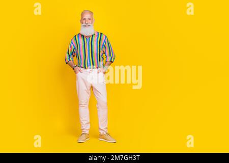 Porträt eines netten, optimistischen alten Mannes mit langem Bart tragen buntes Hemd weiße Hosen Arme in Taschen isoliert auf gelbem Hintergrund Stockfoto