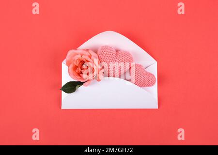 Grußkarte zum Valentinstag, Muttertag und Geburtstag. Rosa Rose und gestrickte rosa Herzen auf rotem Hintergrund in einem Umschlag Stockfoto