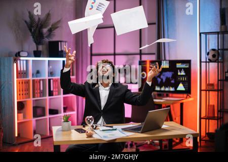 Ein überfreulicher junger, bärtiger Geschäftsmann im Anzug, der gute Nachrichten bekommt, gute Geschäfte macht, Zeitungen in die Luft wirft, feiert und Spaß hat, Überstunden im Abendbüro macht. Stockfoto
