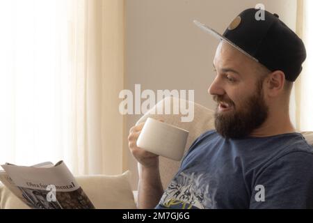 Kaukasischer bärtiger Mann, der Kaffee trinkt und eine Zeitschrift liest. „Obra Fundamental“ bedeutet grundlegende Arbeit. Stockfoto