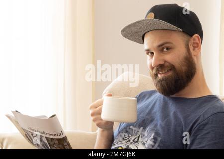 Ein weißer bärtiger Mann, der eine Tasse Kaffee genießt, während er eine Zeitschrift liest. „Obra Fundamental“ bedeutet grundlegende Arbeit. Stockfoto
