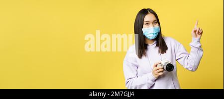 Happy Asian Mädchen in Gesichtsmaske, fotografieren, Zeigefinger auf Kopie Raum, Kamera halten, stehen über gelbem Hintergrund Stockfoto