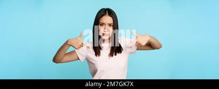 Freche und coole asiatische Frau, die mit den Fingern auf das Logo zeigt und komplizierte, über blauem Hintergrund stehende Augenbrauen mit Smth und Furche zeigt Stockfoto