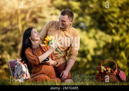 Ein süßes schwangeres Paar, das ein Picknick im Park genießt. Der junge Mann überraschte seine Frau. Stockfoto