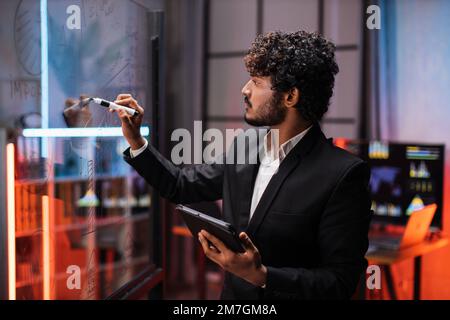Nahaufnahme durch den Glasblick eines fokussierten jungen, bärtigen Geschäftsmanns, der auf einem Glasbrett Kuchendiagramme zeichnet, während er nachts in einem modernen Büro arbeitet. Stockfoto