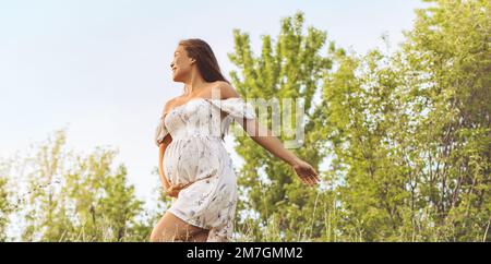 Schwangerschaftsporträt. Schwangere Frau glücklich und lächelnd voller Freude im 3. Trimester in der Natur des Frühlingswaldes. Asiatisches Mädchen, das den Bauch hält Stockfoto
