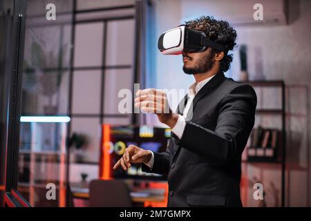 Ein Geschäftsmann mit VR-Brille, der im Büro steht und Gesten mit den Händen macht. Junger männlicher Betriebsangestellter mit Einweisung in moderne Technologie. Menschenbegriff und erweiterte Realität. Stockfoto