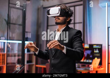 Ein Geschäftsmann mit VR-Brille, der im Büro steht und Gesten mit den Händen macht. Junger männlicher Betriebsangestellter mit Einweisung in moderne Technologie. Menschenbegriff und erweiterte Realität. Stockfoto