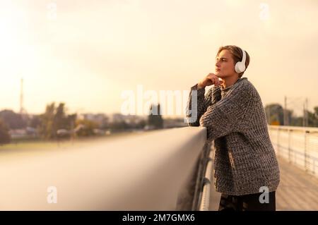 Eine ruhige junge Frau mit kurzen Haaren und Kopfhörern, die auf der Brücke steht und die Stadt bei Sonnenuntergang betrachtet. Stockfoto