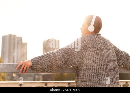 Stilvolle junge Frau mit kurzen Haaren und Kopfhörern, die auf der Brücke steht und die Stadt bei Sonnenuntergang anblickt. Stockfoto