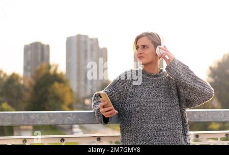 Stilvolle blonde Frau mit kurzen Haaren in Kopfhörern, die ihr Handy in der Hand hält und auf einer Brücke gegen die Stadt steht. Stockfoto