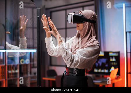 Selbstbewusste muslimische Frau im Hidschab, die innovative Technologie während der Geschäftsstrategie einsetzt. Junge weibliche Mitarbeiter im Unternehmen, die während eines Meetings im Bürozimmer ein VR-Headset tragen. Stockfoto