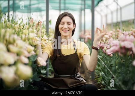 Hübsche Frauen, die sich unter Blumen entspannen, während sie in einer Meditationshaltung im grünen Haus sitzen Stockfoto