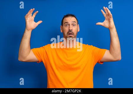 Hispanischer Mann träumt bärtig melancholisch überraschend attraktiver Mann 40s in lässigem orangefarbenem Basis-T-Shirt verteilt Hände isoliert auf blauem Hintergrund Stockfoto