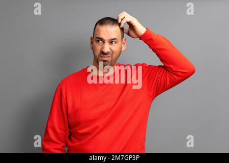 Bärtiger hispanischer Mann in rotem Pullover, runzelnd und unzufrieden, während er sich am Kopf kratzt, über blauem Hintergrund Stockfoto