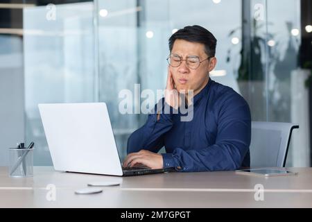 Ein junger Asiate arbeitet im Büro an einem Schreibtisch mit einem Laptop. Er hält seine Wange. Er hat starke Zahnschmerzen, er braucht die Hilfe eines Zahnarztes. Stockfoto