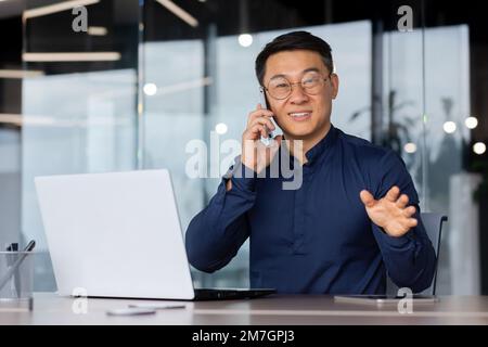 Ein junger Asiate telefoniert im Büro. Er sitzt mit einem Laptop am Schreibtisch, sieht in die Kamera und lächelt. Stockfoto