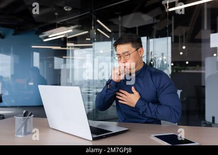 Ein junger Asiate hustet im Büro am Schreibtisch. Hat Allergien, Grippe, Asthma. Er hält seine Brust, bedeckt seinen Mund. Stockfoto