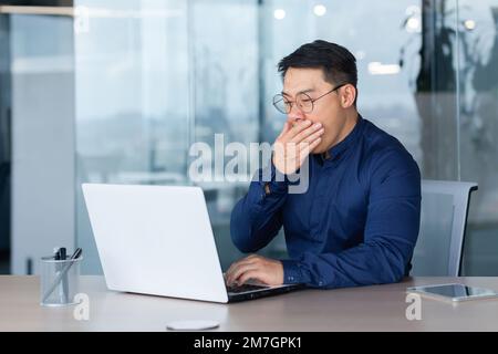 Müder junger asiatischer Geschäftsmann gähnt am Arbeitsplatz. Er sitzt schläfrig am Schreibtisch im Büro und arbeitet Überstunden am Laptop. Stockfoto