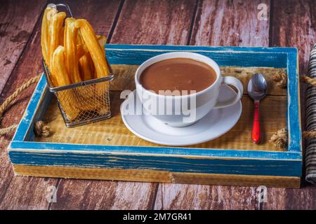 Heiße Schokolade in einem weißen Becher mit Churros in einem blauen Holztablett auf einem Holztisch Stockfoto