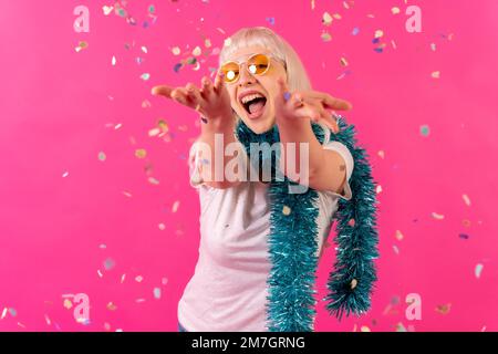 Lächelnd auf einer Party, mit Konfetti in Sonnenbrille, blondes, weißhäutiges Mädchen im rosa Studio Stockfoto