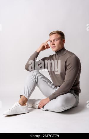 Porträt eines attraktiven blonden kaukasischen Modells in einem braunen Pullover auf weißem Hintergrund, sitzend Stockfoto