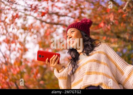 Porträt einer Asiatin im Herbst mit einem Handy, das eine Sprachnachricht in einem Wald aus roten Blättern sendet, Technologie Stockfoto
