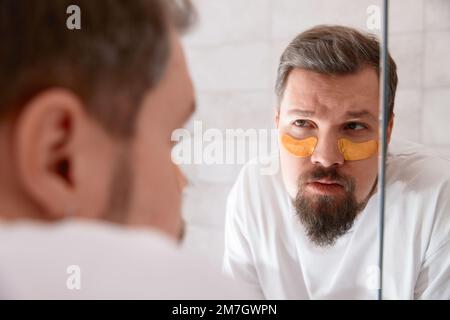 Porträt eines Mannes in weißem T-Shirt, der im Badezimmer Augenflecken auf sein Gesicht aufträgt. Self-Care-Verfahren am Morgen Stockfoto