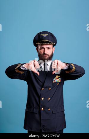 Seriöser Flugzeugpilot mit Daumen-nach-unten-Geste trägt professionelles, einheitliches Porträt, Flugzeugkapitän schaut auf die Kamera. Der Pilot zeigt ein Missbilligungszeichen mit Fingern, Studio-Medium-Shot Stockfoto