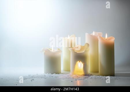 Eine Gruppe weißer Kerzen, eine brennt in einer warmen Höhle, surreales Stillleben als Energiesparkonzept, verschwenden Sie nicht die Hitze draußen, hellgrau Stockfoto