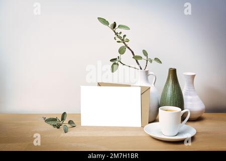 Modell einer leeren weißen Grußkarte mit einem naturbraunen Umschlag, drei Vasen mit einem Salbeiblatt-Zweig und einer Kaffeetasse auf einem Holztisch vor einem Stockfoto