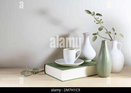 Noch immer leben von drei Vasen mit einem Salbeiblatt-Zweig, einem grauen grünen Buch und einer Kaffeetasse auf einem Holztisch oder Tisch vor einer hellgrauen Wand, ruhiges Zuhause Stockfoto