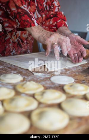 Nicht wiedererkennbare alte Frau schneidet Blätterteig mit einem Glas, um Knödel zuzubereiten. Spanien Stockfoto