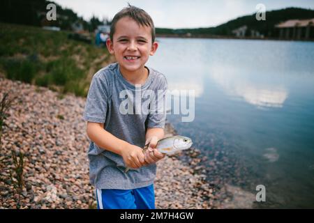 Der Junge lächelt mit Fisch, den er im Urlaub im Bergsee gefangen hat Stockfoto