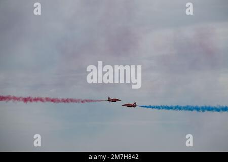 Zwei Flugzeuge der Serie BAE Systems Hawk T1 mit roten Pfeilen führen ein Kreuzmanöver durch und führen roten und blauen Rauch auf einer Ausstellung über Eastbourne, East Sussex, durch Stockfoto